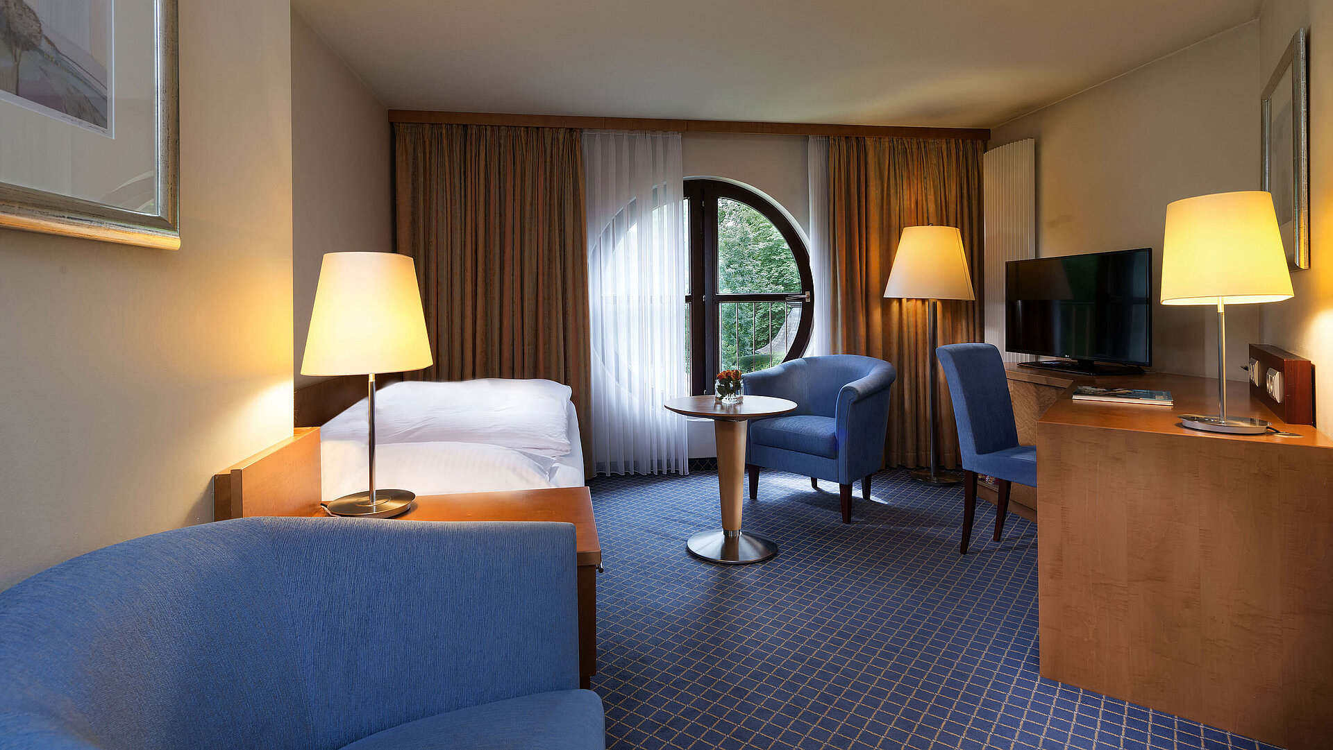 Habitación Classic en Maritim Hotel Fulda con cama individual, escritorio, zona de estar y ventana redonda con vistas.