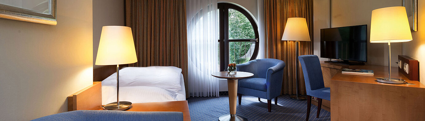 Chambre Classic au Maritim Hotel Fulda avec lit simple, bureau, coin salon et fenêtre ronde avec vue.