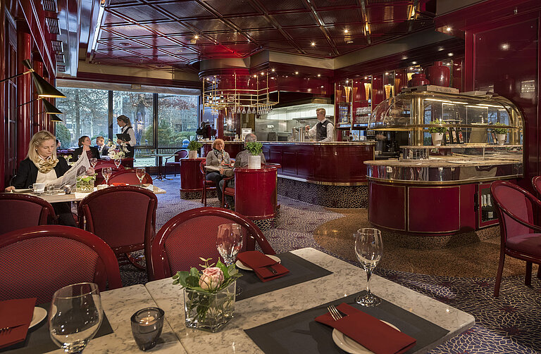 The image shows the cozy "Brasserie" café at the Maritim Hotel Bonn with red decor and a relaxed atmosphere.