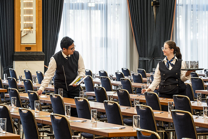 Miembros del personal en el Maritim Hotel Magdeburg preparando el salón con vasos y materiales en las mesas.