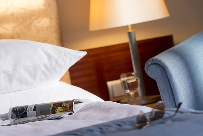 Primer plano de una habitación en el Maritim Hotel Fulda con cama, revista, gafas y un vaso de agua en la mesita de noche.