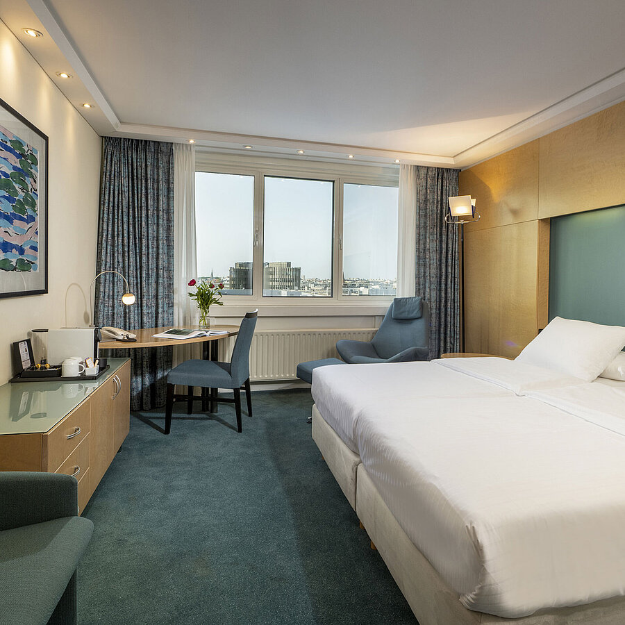 Chambre Supérieure meublée moderne à l'hôtel Maritim avec grand lit, bureau,œuvre d'art et vue sur la ville depuis la fenêtre