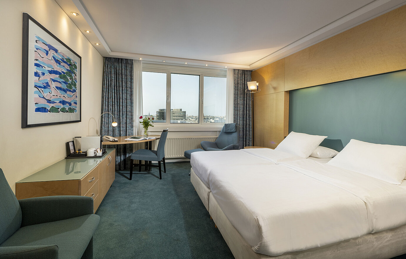 Chambre Supérieure meublée moderne à l'hôtel Maritim avec grand lit, bureau,œuvre d'art et vue sur la ville depuis la fenêtre