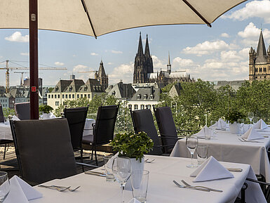 Terraza en la azotea con vistas al Rin y al puente Hohenzollern, con mesas puestas bajo sombrillas.