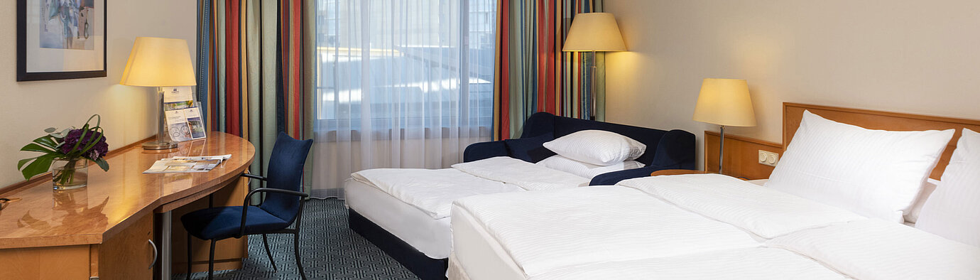 Chambre familiale Classic du Maritim Hotel Frankfurt avec lit double, canapé-lit, bureau et rideaux colorés.