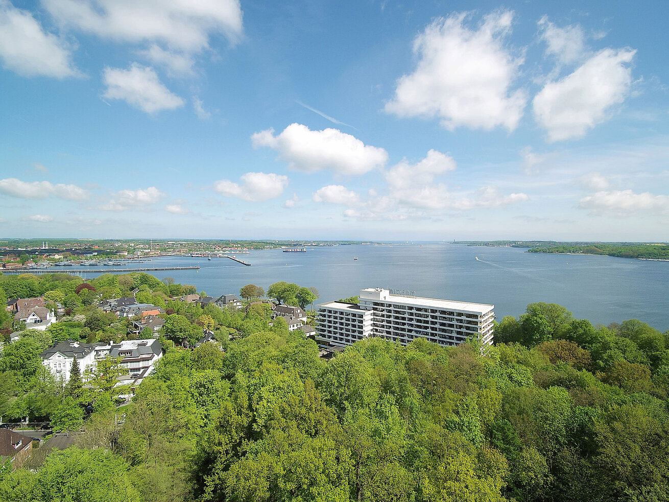 Vista aérea del Maritim Hotel Kiel, rodeado de verdes paisajes y el fiordo de Kiel, bajo un cielo azul brillante.