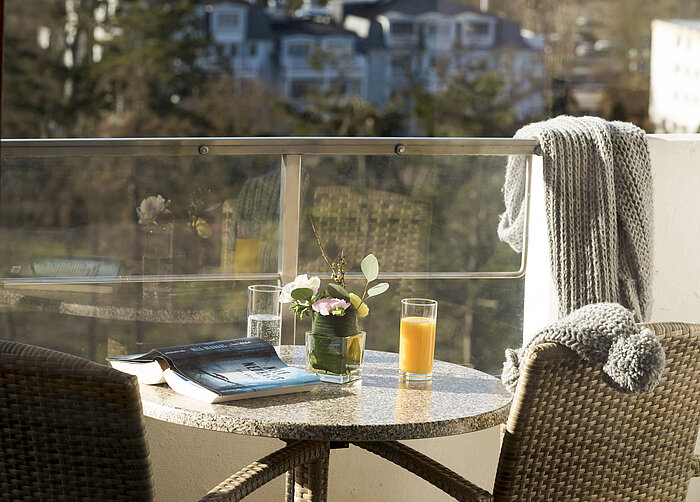 Table de balcon avec boissons, livre et fleurs au Maritim Seehotel Timmendorfer Strand avec vue sur arbres et bâtiments.