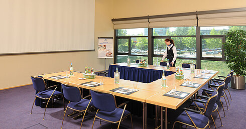 Sala de reuniones del Maritim Hotel Dresde con mesas en U, pantalla de presentación y preparativos para una reunión.