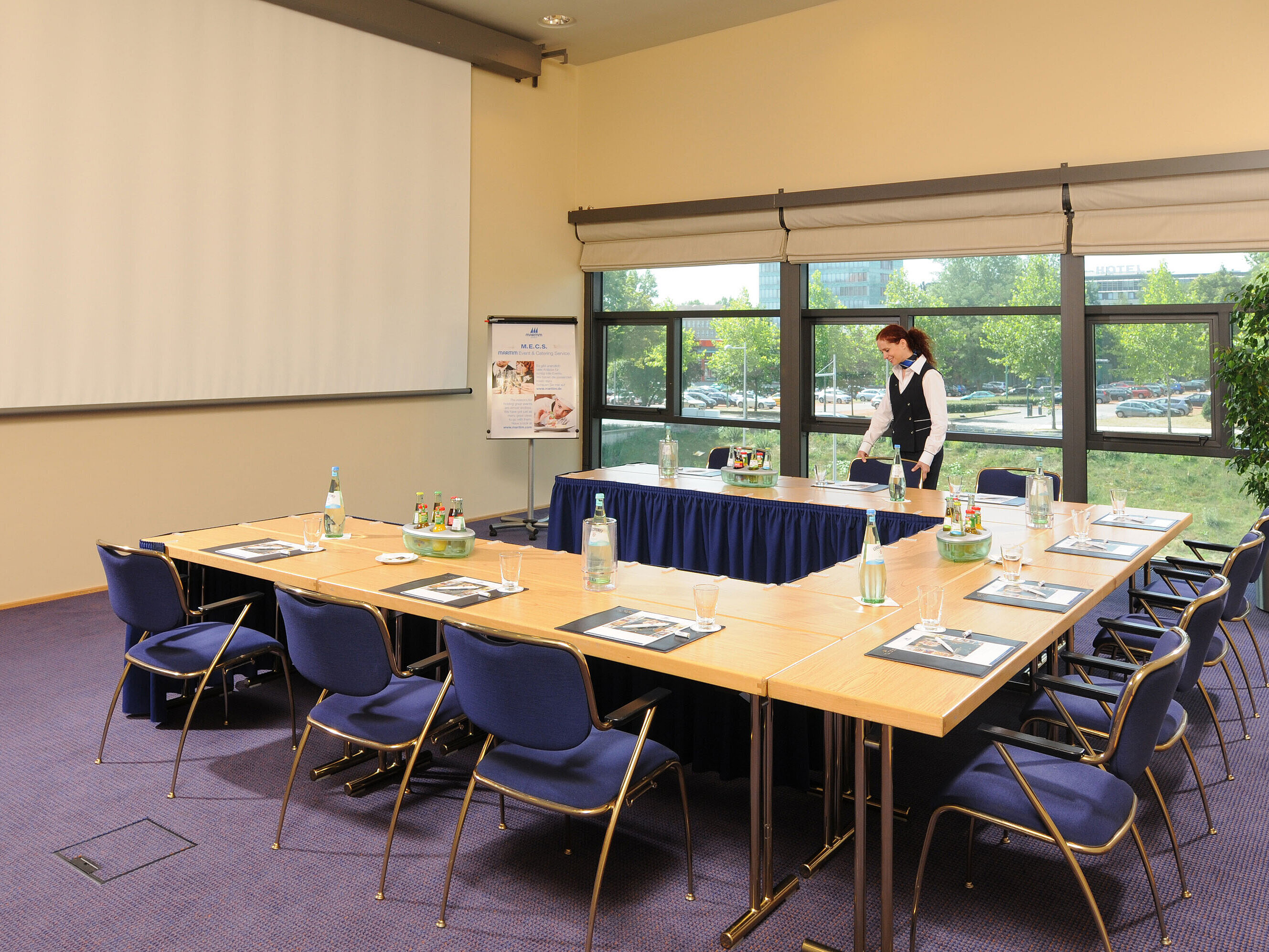 Sala de reuniones del Maritim Hotel Dresde con mesas en U, pantalla de presentación y preparativos para una reunión.