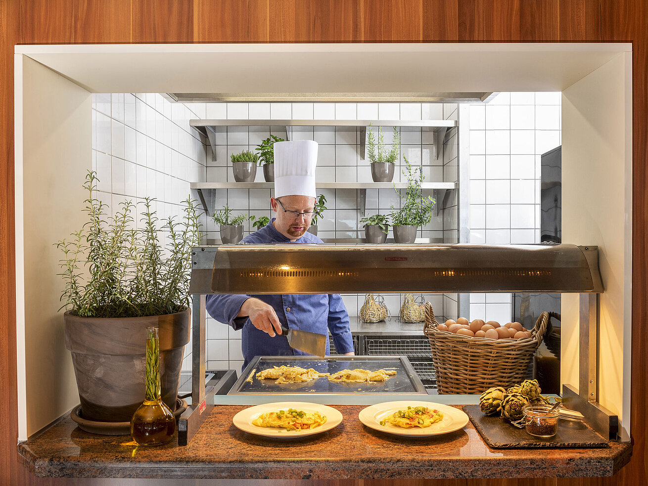 Un chef prépare des omelettes à une station de cuisson entourée d'herbes fraîches. Deux assiettes de nourriture sont prêtes.