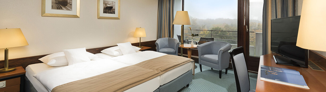 Habitación clásica del Maritim Hotel Kiel con cama doble grande, zona de estar, escritorio y balcón con vistas a la vegetación.