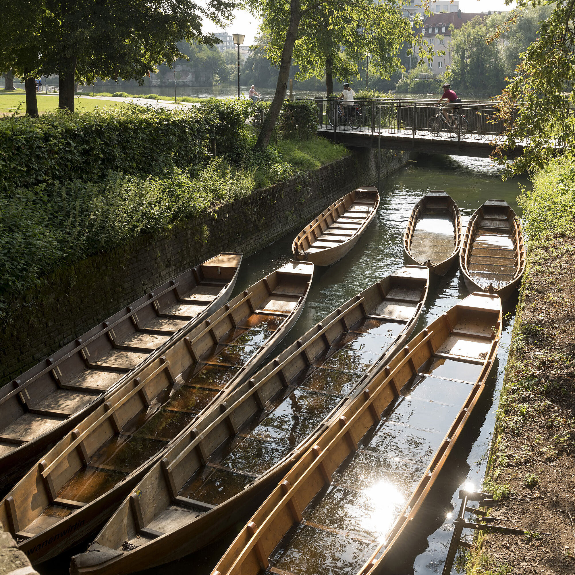 Wooden boats in a side channel of the Danube in Ulm, surrounded by green trees.