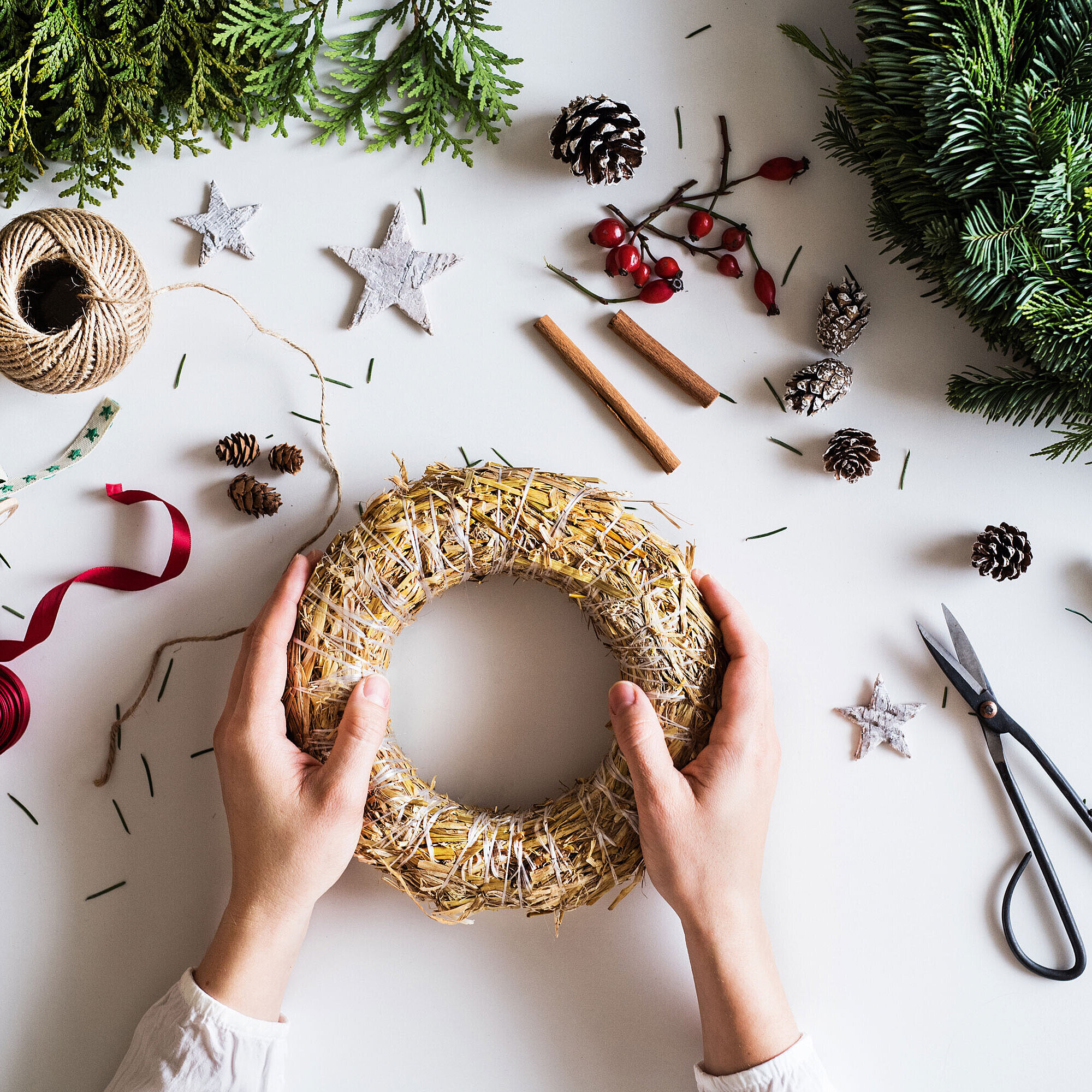 Hands arranging an Advent wreath on a table with fir branches, ribbons and craft materials.