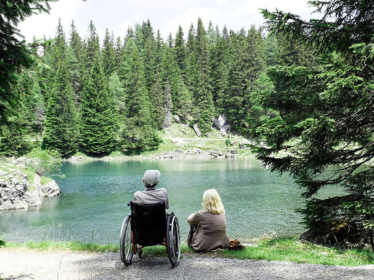 © Herby Meseritsch – AdobeStock.com Dos personas, una en silla de ruedas, sentadas junto a un lago mirando el bosque