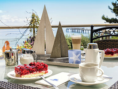 Table avec gâteau et boissons chaudes sur la terrasse du Maritim Seehotel Timmendorfer Strand avec vue sur la mer.