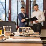 Conference room with a laptop, drinks, and documents, two businessmen in the background discussing papers.
