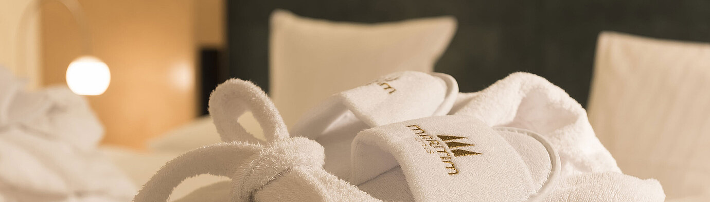 Accappatoio bianco e pantofole con logo Maritim sul letto, ideali per momenti di relax nella camera d’hotel