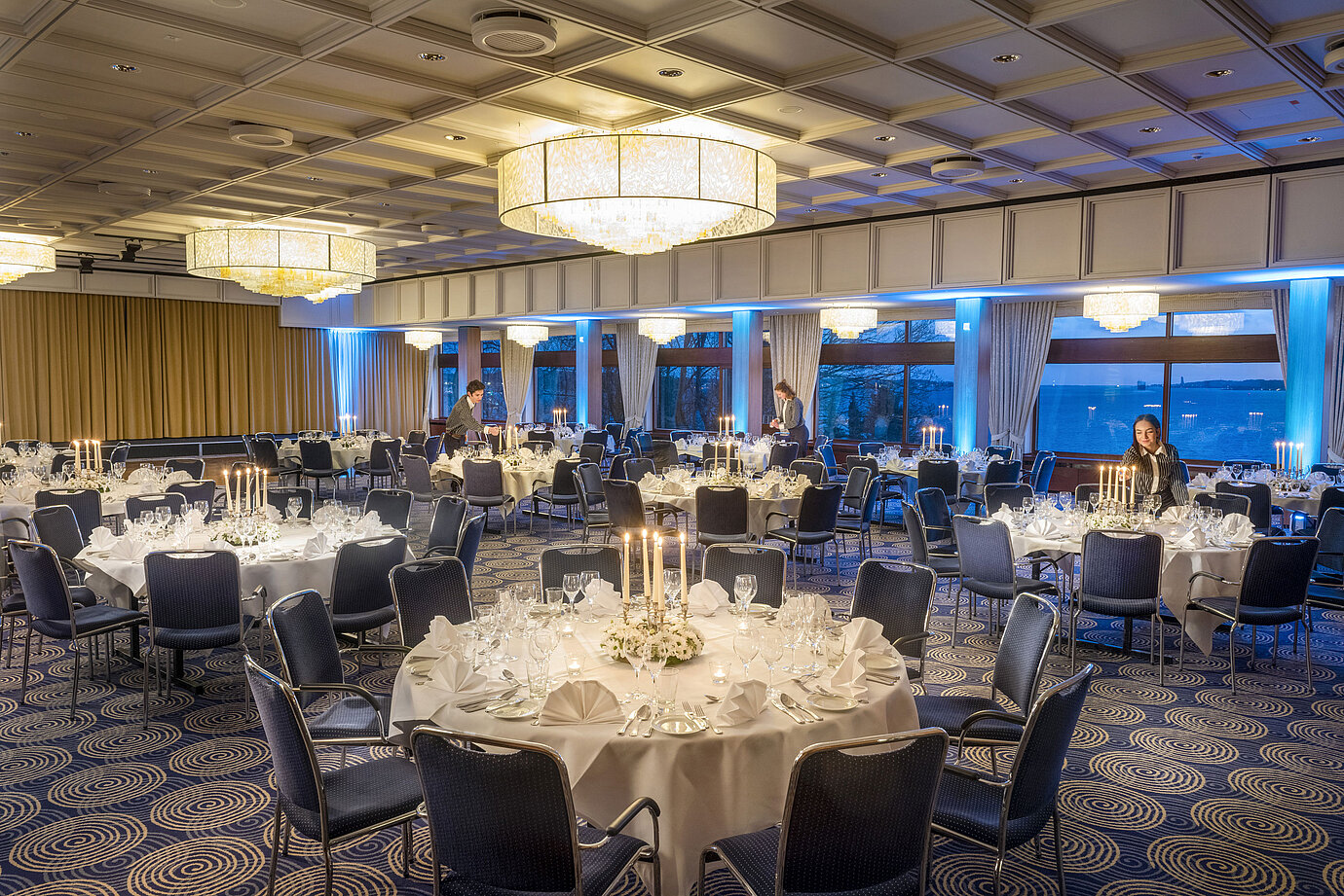 Sala Maritim del Hotel Maritim de Kiel, mesas dispuestas festivamente a la luz de las velas, ambiente elegante, personal preparando el evento.