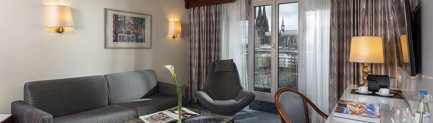 Suite Junior Suite Junior à l'Hôtel Maritim Cologne avec vue sur la cathédrale de Cologne, canapé élégant, fauteuil et bureau avec lecture