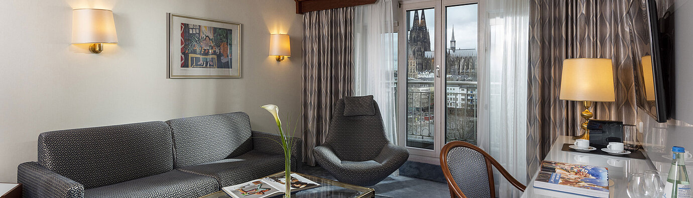 Suite Junior à l'Hôtel Maritim Cologne avec vue sur la cathédrale de Cologne, canapé élégant, fauteuil et bureau avec lecture