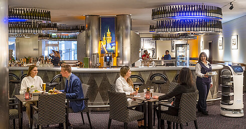 Des clients dînent au bar de l'hôtel Maritim proArte Berlin, entourés de services modernes et d'un robot serveur.