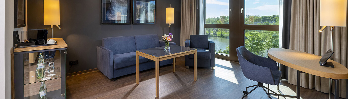 Suite Junior Suite lumineuse avec coin salon, grande fenêtre et vue sur la nature à l'hôtel Maritim Ingolstadt.