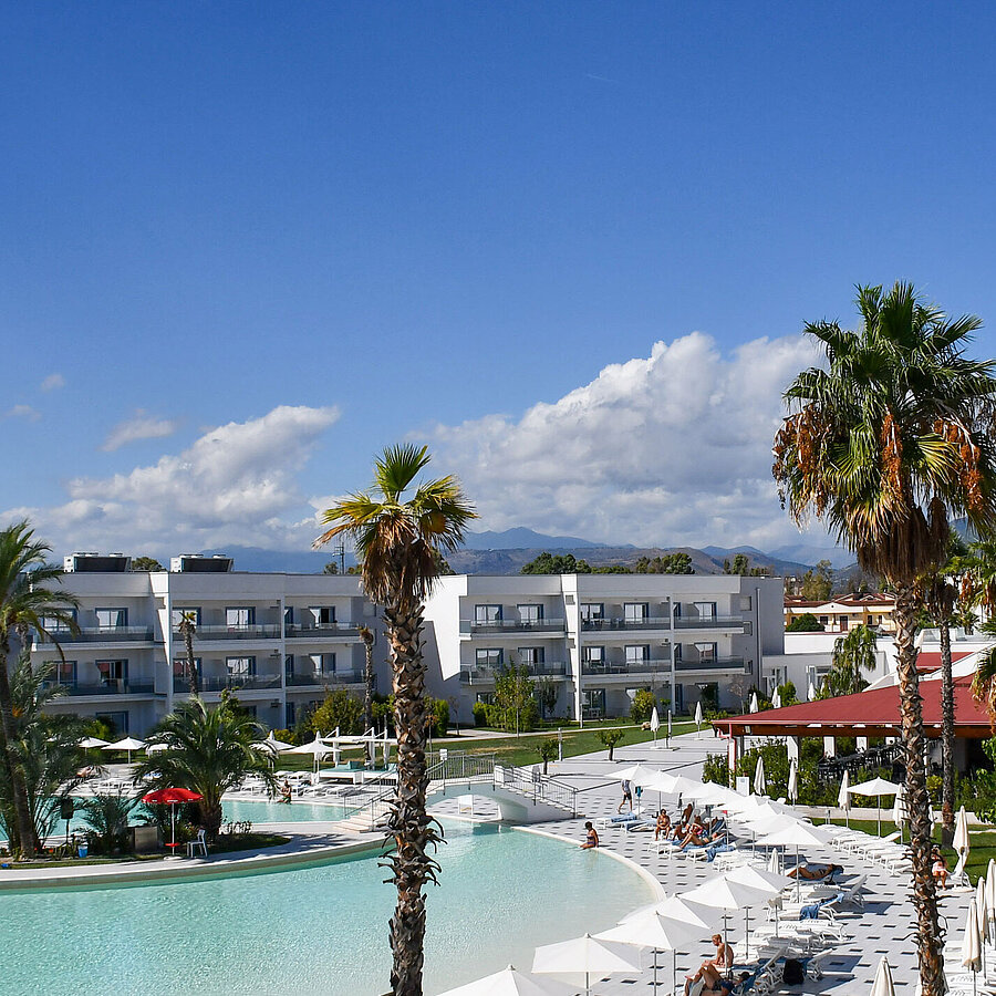Exterior view Panorama of Maritim Resort Calabria with pool, palm trees, white buildings, and mountain backdrop under clear skies.