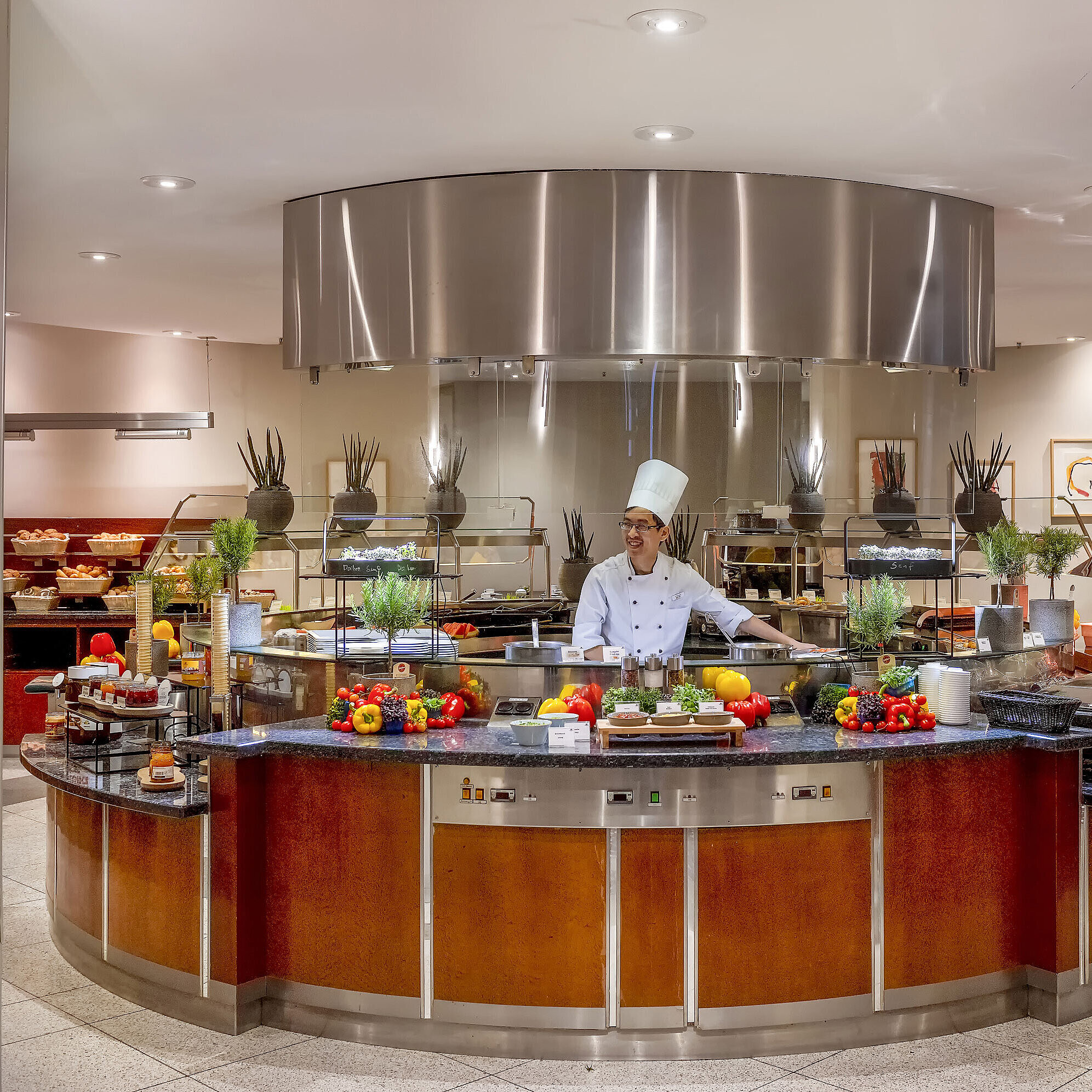 Front cooking Chef at the show cooking station at Maritim proArte Hotel Berlin, surrounded by fresh vegetables and herbs.